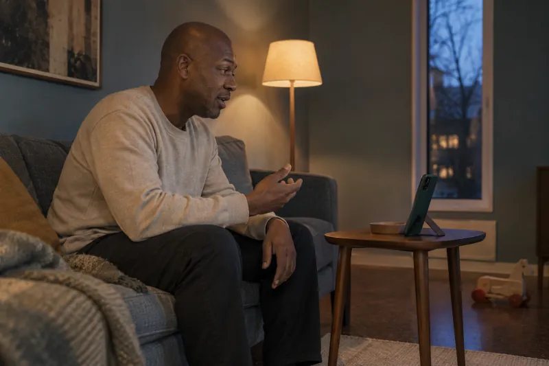 A parent practises speaking toward a propped-up phone in a softly lit living room at dusk.