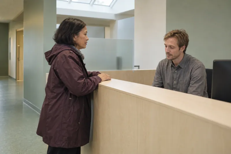 An adult talks calmly with a receptionist at a health-centre counter.